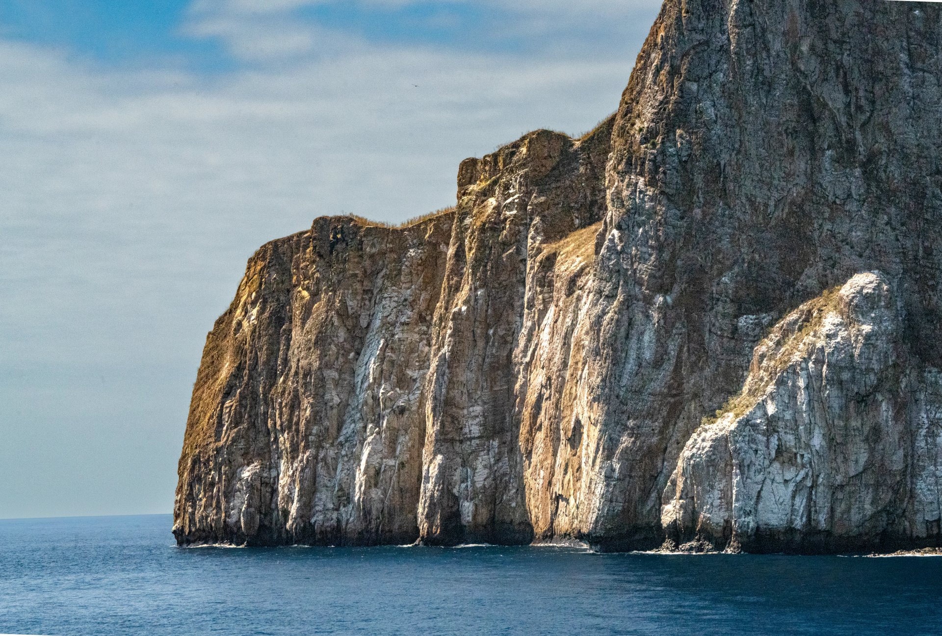 A close-up fine art photograph of a towering, heavily textured volcanic cliff face rising out of blue ocean water in the Galapagos Islands, bathed in warm sunlight.