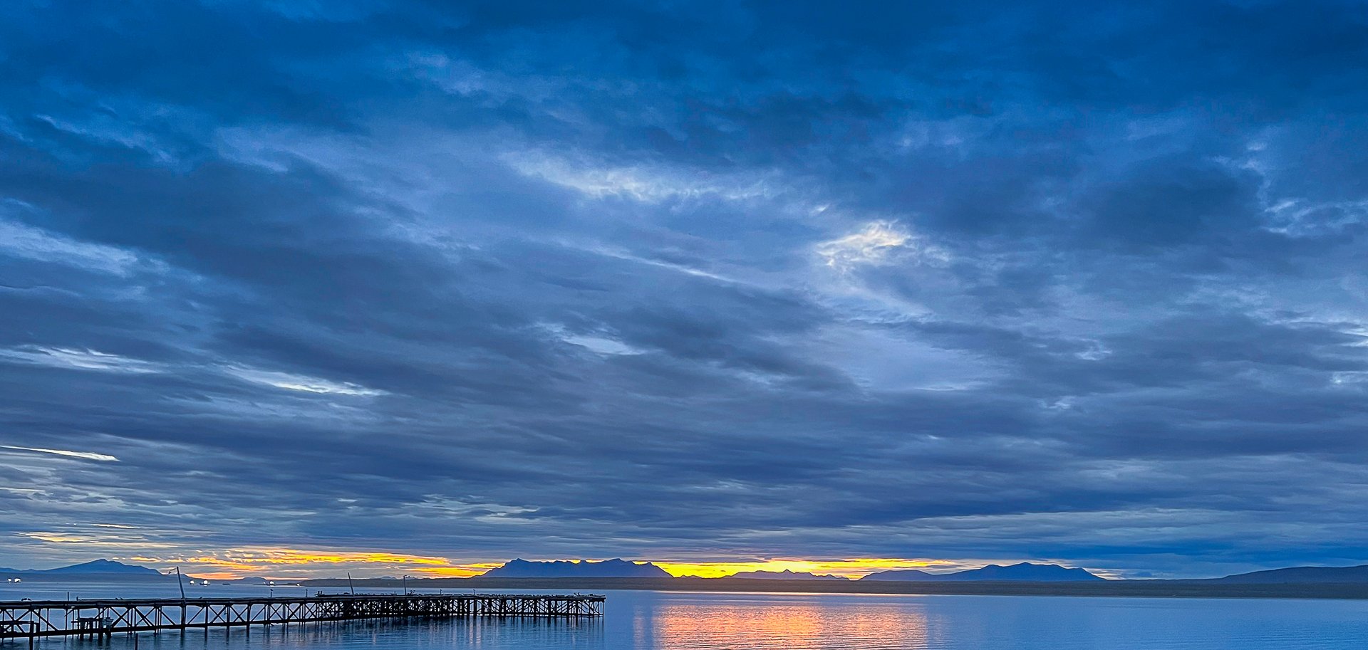 Scenic sunset over calm waters with a wooden pier and mountains in the background.